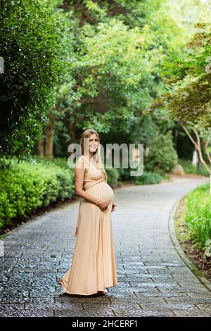 Schwangere Frau in Abendkleid hält Hände auf Bauch im Freien in Bambuswald. Schwangerschaft, Mutterschaft, Vorbereitung und Erwartung Konzept. Wunderschön Stockfoto