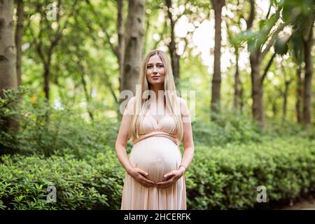 Schwangere Frau in Abendkleid hält Hände auf Bauch im Freien in Bambuswald. Schwangerschaft, Mutterschaft, Vorbereitung und Erwartung Konzept. Wunderschön Stockfoto