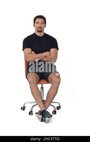 Vorderansicht eines Vollportraits eines Mannes, der auf einem Stuhl sitzt, gekreuzte Arme, Sportkleidung auf weißem Hintergrund tragend Stockfoto