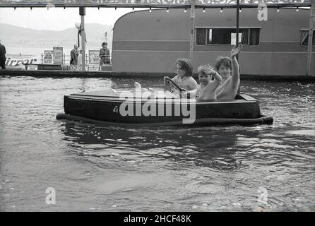 1960s, geschichtlich, drei kleine Kinder in einem kleinen hölzernen Motorboot auf einem See, der am Meer Boot fährt, der kleine Junge und eines der Mädchen, die beide am Lenkrad festhalten, England, Großbritannien. Mit einem „Stoßfänger“ aus Gummi um den Rand wurden diese kleinen Elektroboote speziell für den Einsatz in Binnengewässern an der Küste entwickelt. Ein Vorläufer war die „Glida“, ein kleines Boot, das 1929 erstmals auf dem Bootssee im Battersea Park, London, eingesetzt wurde. Stockfoto