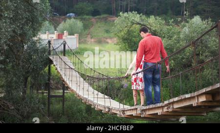 Junge Liebhaber auf einer Seilbrücke über den Fluss. Ein paar Wanderer überqueren eine Schaukelbrücke. Clip. Junges Paar in der Liebe zu Fuß auf schwebenden Holz b Stockfoto