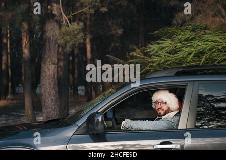 Mann mit Weihnachtsmannhut im Auto mit Tannenbaum auf der Oberseite. Weihnachten, Winterferien im Zusammenhang mit einem kaukasischen Männchen Stockfoto