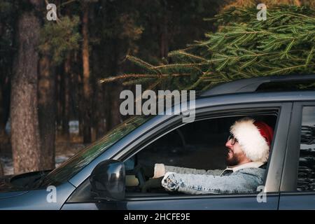 Mann mit Weihnachtsmannhut im Auto mit Tannenbaum auf der Oberseite. Weihnachten, Winterferien im Zusammenhang mit einem kaukasischen Männchen Stockfoto