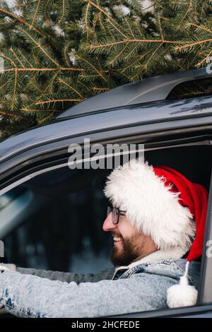 Mann mit Weihnachtsmannhut im Auto, Weihnachtsbaum auf dem Dach. Neues Jahr, Winterferien verwandte Konzept, glücklich zuversichtlich kaukasischen Porträt Stockfoto