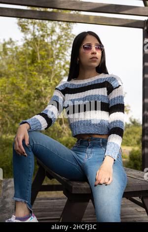 Posiert junge latinerin mit geradem schwarzen Haar, entspannt auf einer Parkbank sitzend, Sonnenbrille und lässige Kleidung, Schönheit und Mode tragend Stockfoto
