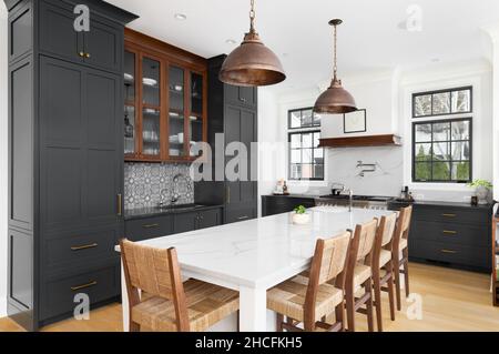 Eine luxuriöse Küche mit dunkelgrauen Schränken, bronzefarbenen Deckenleuchten über der großen weißen Insel und luxuriösen Geräten. Stockfoto