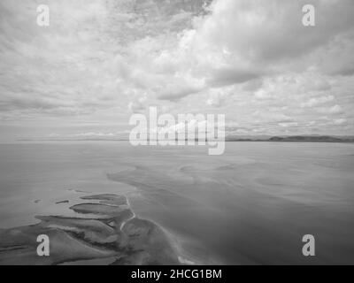 Schwarz-weißes Bild von abstrakten Sandmustern bei Ebbe auf dem Ozean, das sich mit einem bewölkten Himmel und einem fernen Horizont vermischt Stockfoto