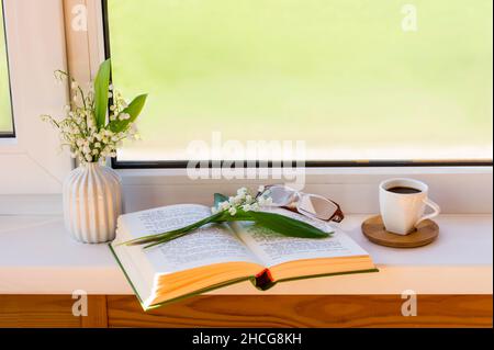 Ein offenes Buch liegt auf der Fensterbank mit einem Bouquet von Frühlingslilien des Tales Stockfoto