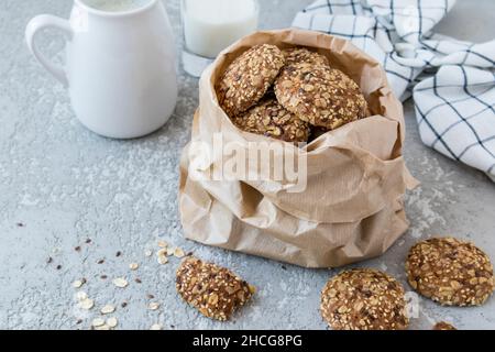 Haferflocken-Kekse in einem Bastelsack auf einem Betonhintergrund mit einem Krug Milch und einem gegossenen Glas Milch. Nahaufnahme Stockfoto