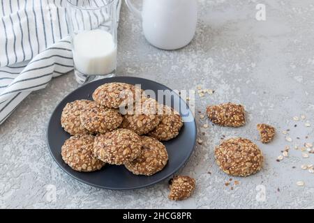 Haferflockenkekse mit Leinsamen, Sesam und Rosinen auf einem dunklen Teller und einem Glas Milch auf grauem Grund Stockfoto