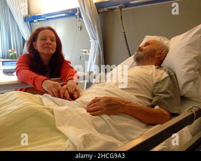 83-jähriger Mann, der in einem Krankenhausbett liegt. Seine Tochter sitzt neben dem Bett und hält seine Hand. Stockfoto