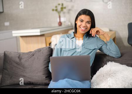 Junge Freiberuflerin mit gemischter Rasse und brünettem Haar in bequemer Freizeitkleidung, die auf der Couch sitzt und an dem Projekt arbeitet, E-Mails auf dem Laptop tippt und einen Artikel schreibt Stockfoto