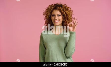 Glückliche Rothaarige Frau im grünen Jumper zeigt okay Geste isoliert auf rosa Stockfoto