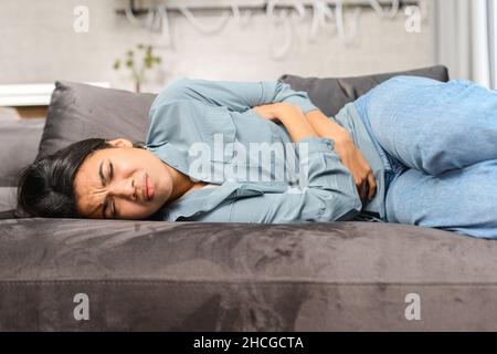 Traurige junge multirassische Frau, die unter Menstruationsschmerzen leidende Freizeitkleidung trägt, sich am Bauch krank fühlt, den Bauch hält, während der Periode Bauchkrämpfe hat und zu Hause auf dem Bett liegt Stockfoto