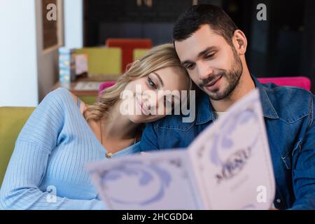 Fröhliches Paar, das Kopf an Kopf sitzt, während es sich aus einem verschwommenen Menü eine Mahlzeit auswählt Stockfoto