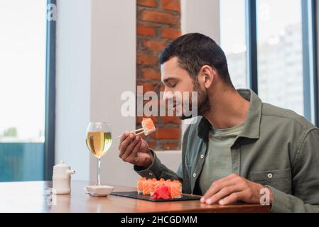 Junger Mann mit geschlossenen Augen hält köstliches Sushi mit Essstäbchen in der Nähe von Glas oder Weißwein Stockfoto