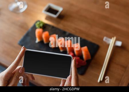 Teilansicht einer Frau, die auf dem Mobiltelefon ein Foto von köstlichem Sushi macht, unscharfer Hintergrund Stockfoto