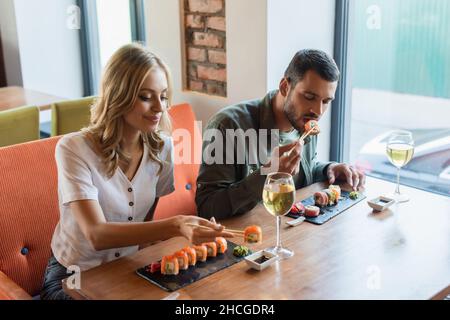 Glückliche Frau, die neben ihrem Freund leckere Sushi-Brötchen und ein Glas Weißwein isst Stockfoto