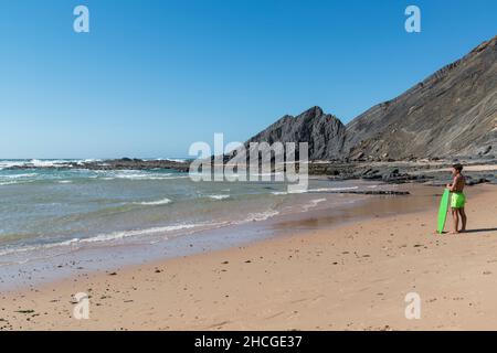 Junger Mann in fluoreszierenden grünen Badehosen mit einem Surfbrett der gleichen Farbe blickt auf das Meer am Strand Praia da Amoreira an der Algarve, Portugal. Stockfoto