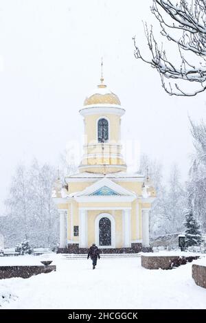 Winter Schneefall in der Stadt. Stadtlandschaft. Christliche Kapelle unter dem Schnee in der Stadt. Eine Frau geht unter dem Schnee zur Kirche. Blizzard. Stockfoto