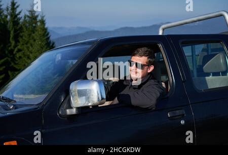 Sicherer Mann mit einem Lächeln auf dem Gesicht in schwarzer Sonnenbrille hinter dem Steuer eines SUV, der seinen Ellbogen an die Seitentür lehnte. Grüne Bäume und Berghügel im Hintergrund. Stockfoto