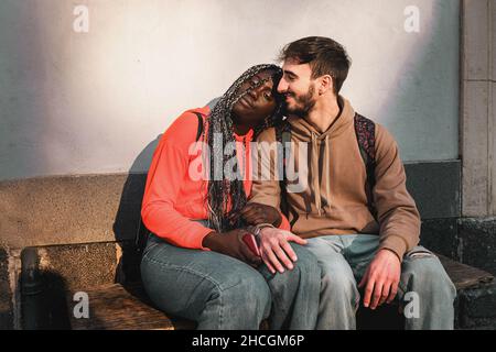 Ein paar junge Menschen, die verschiedene Nationalitäten und Kulturen lieben, ruhen auf der Straße und sitzen auf einer Bank Stockfoto