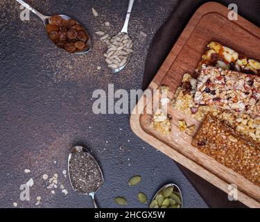 Protein-Müsliriegel. Verschiedene Müsliriegel mit Nüssen, Samen, Hafer, Beeren, trockenen Früchten und Löffeln mit Zutaten auf einem dunkelbraunen Bac Stockfoto