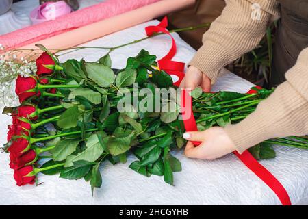 Weibliche Floristin, die einen Strauß roter Rosen macht. Die Hände der kaukasischen Frau binden einen Strauß mit einem roten Band. Draufsicht Stockfoto