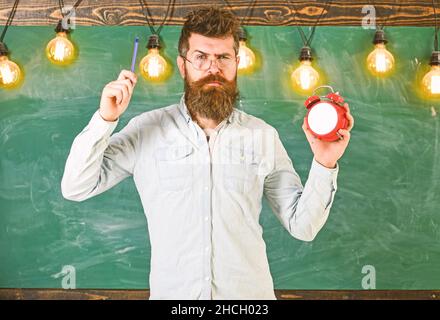 Der Lehrer in der Brille hält Wecker und Stift. Disziplinkonzept. Bärtiger Hipster hält die Uhr, Kreidetafel auf dem Hintergrund. Mann mit Bart und Stockfoto