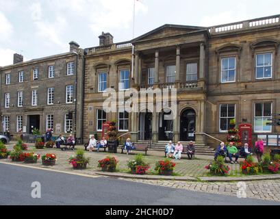 Senioren, die die Sonne genießen, sitzen auf Bänken vor dem Rathaus von Skipton, North Yorkshire, England, mit Blumen in den Wannen im Vordergrund. Stockfoto