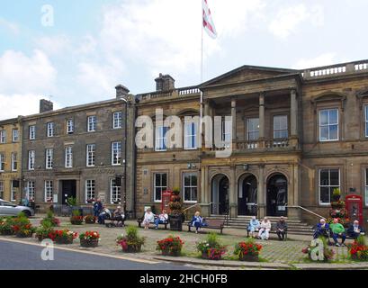 Senioren, die die Sonne genießen, sitzen auf Bänken vor dem Rathaus von Skipton, North Yorkshire, England, mit Blumen in den Wannen im Vordergrund. Stockfoto