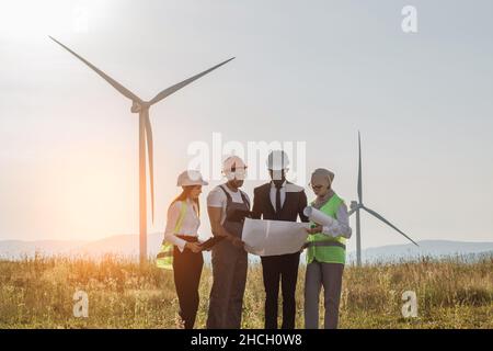 Eine multiethnische Gruppe von Ingenieuren, Technikern und Managern in Schutzhelmen, die sich im Freien über grüne Energie treffen. Männer und Frauen, die auf einem Windmühlenbauernhof mit Tablet und Zeichnung arbeiten. Stockfoto