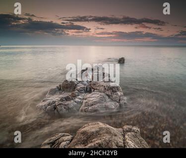 Sonnenuntergang über dem Meer, mit einem ruhigen, seidigen Meer, das um Felsen an der Küste wirbelt. Kieselsteine können durch das klare Wasser gesehen werden. Stockfoto