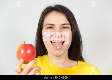 Eine Frau hält in ihrer Hand einen Apfel, eine weiße Tablette mit Vitaminen oder Nahrungsergänzungsmitteln auf der Zunge. Stockfoto