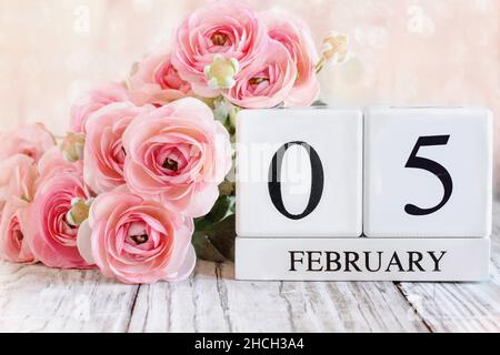 Weiße Holz-Kalenderblöcke mit dem Datum Februar 5th und rosa Ranunculus-Blumen über einem Holztisch. Selektiver Fokus mit unscharfem Hintergrund. Stockfoto