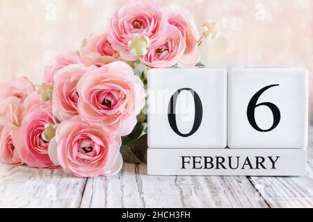 Weiße Holz-Kalenderblöcke mit dem Datum Februar 6th und rosa Ranunculus-Blumen über einem Holztisch. Selektiver Fokus mit unscharfem Hintergrund. Stockfoto