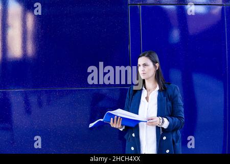 Schöne kaukasische Geschäftsfrau, die Dokumente in einem Ordner vor einem Bürogebäude liest. Sie trägt eine lässige blaue Jacke. Stockfoto