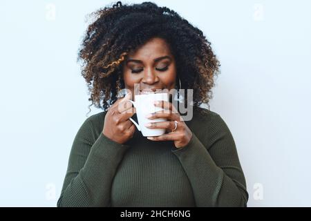 Porträt einer lächelnden afroamerikanischen schwarzen Frau mit mittleren Erwachsenen, die Kaffee trinkt und die Augen vor weißem Hintergrund geschlossen hat Stockfoto