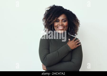 Schöne Frau mittleren Alters schwarz afroamerikanisch lächelnd mit lockigen Haaren trägt einen grünen Pullover mit gekreuzten Armen Stockfoto