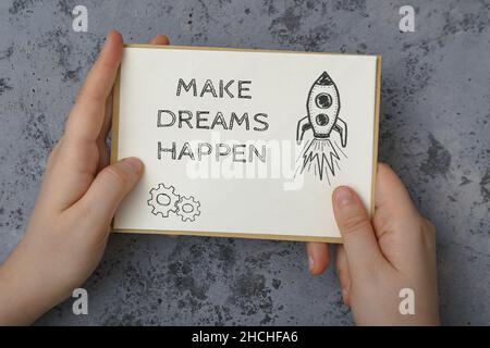 Das Mädchen hält einen Brief in ihren Händen mit dem Text Make Dreams Happen. Motivierendes Zitat auf einem weißen Blatt Papier. Stockfoto