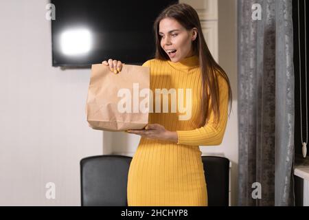 Ein junges kaukasisches Mädchen steht allein zu Hause und hält eine Papiertüte mit Essen in den Händen. Konzept der Bestellung Lieferung von Lebensmitteln nach Hause oder zum Mitnehmen. Stockfoto