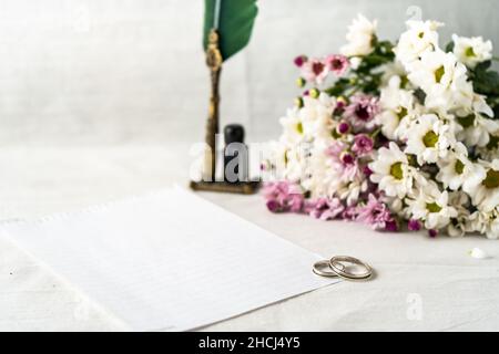 Einige Hochzeit oder Verlobungsringe zusammen mit einer alten Federkiel in seinem Tintenfass und einem weißen Blatt bereit, eine Notiz zu schreiben. Schöne Blumen begleiten den Compo Stockfoto