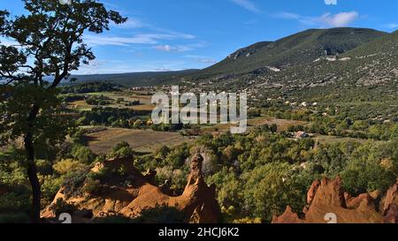 Schöne Aussicht auf das Luberon-Tal mit bunten ockerfarbenen Felsen bei Colorado Provenzal mit Rustrel-Dorf im Hintergrund in der Provence, Frankreich. Stockfoto