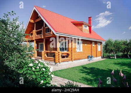 Großes neu gebautes Holzhaus mit rotem Ziegeldach, Rasenfläche auf der rechten Seite und Kirschbaumgarten hinter dem Haus und vor dem Blumengarten Stockfoto