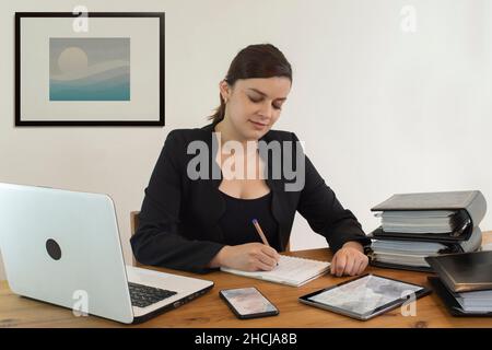 Schöne hispanische Angestellte, die glücklich lächelte und an ihrem Schreibtisch saß und mit dem Schreiben in ihrem Notebook gegen die weiße Wand beschäftigt war Stockfoto