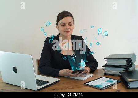 Schöne hispanische Geschäftsführerin, die glücklich lächelt und an ihrem Schreibtisch sitzt und eine E-Mail von ihrem Smartphone mit Mail-Symbolen verschicken lässt Stockfoto