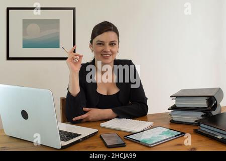 Wunderschöne hispanische Angestellte im Büro mit einer lächelnden Brille, die mit einem Computer und einem Smartphone am Schreibtisch saß und sich nach vorne freute Stockfoto