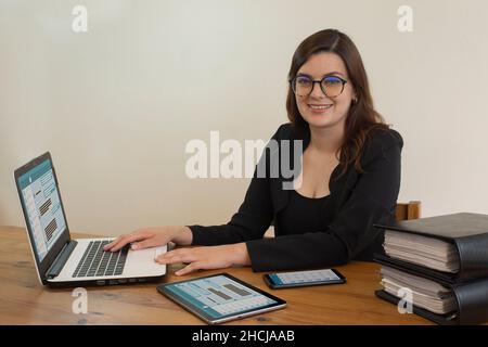 Wunderschöne hispanische Büroangestellte mit einer lächelnden Brille, die vor ihrem Computer vor der weißen Wand sitzt und fröhlich an ihrem Schreibtisch sitzt Stockfoto