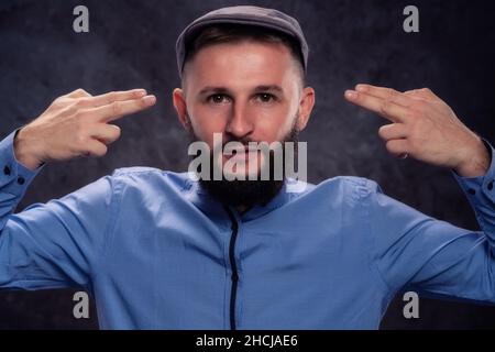 Der junge bärtige Europäer mit einer Mütze imitiert mit seinen Fingern einen Kopfschuss. Für Werbung auf dunklem Hintergrund Stockfoto