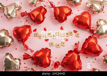 Das Wort Valentinstag aus Buchstabenblöcken und roten Herzen Foil Ballons herum. Romantisches Konzept zum Valentinstag. Draufsicht. Stockfoto